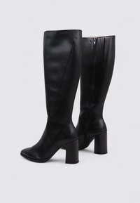 Botas negras hasta la rodilla, hechas de cuero suave, con puntera cuadrada y tacón grueso. Presentan un cierre lateral y un diseño elegante.