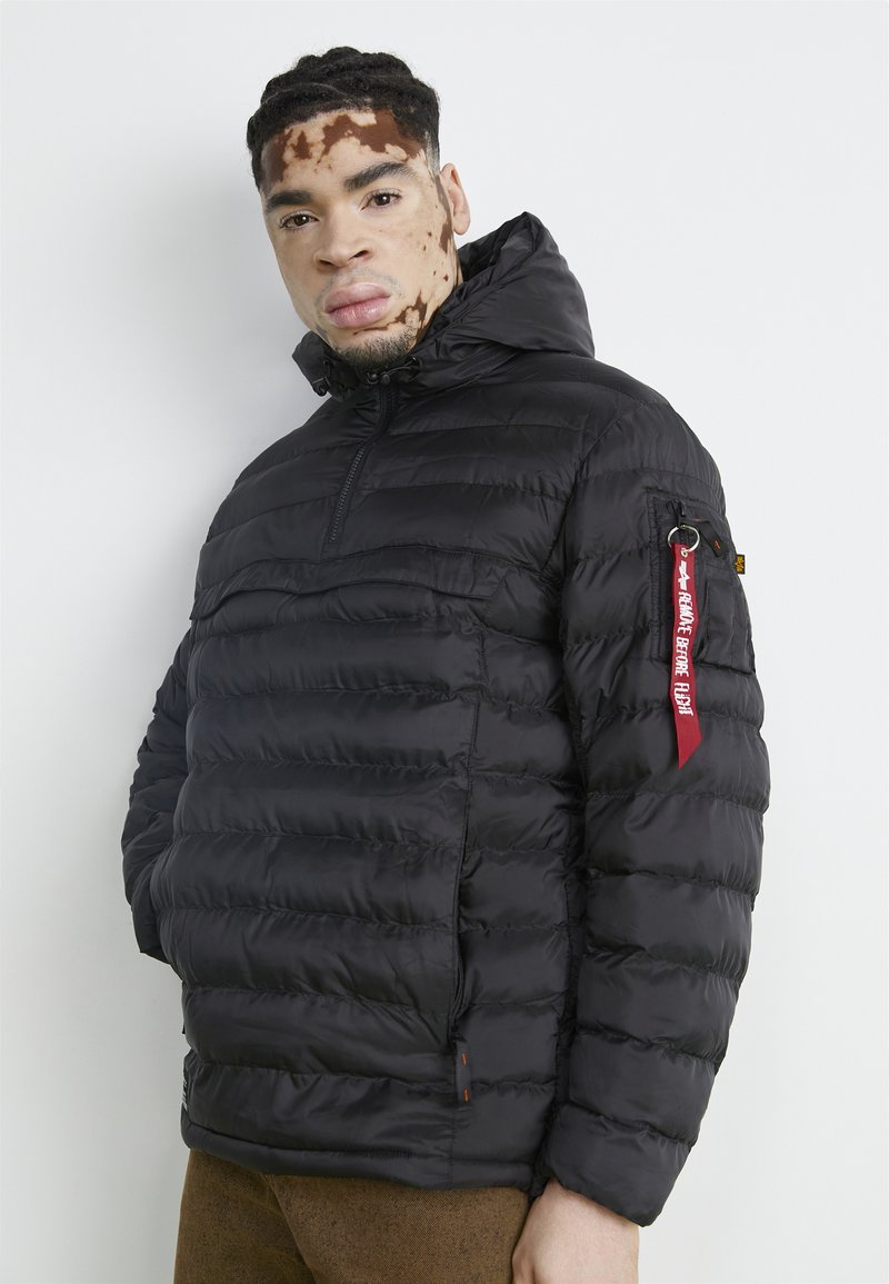 Giubbotto trapuntato nero con design a rombi, cappuccio, chiusura lampo frontale e un'etichetta in tessuto rossa sulla manica sinistra. Texture morbida e liscia.