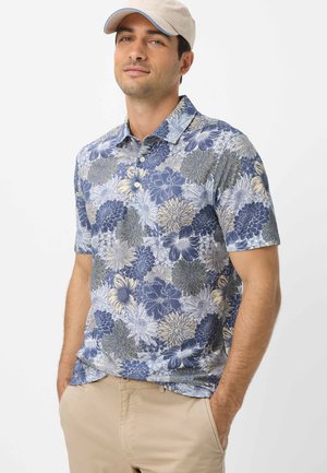 Mann trägt beige Kappe, blaues Polohemd mit floralem Muster und kurzen Ärmeln sowie beige Hose, steht mit den Händen in den Taschen vor weißem Hintergrund.