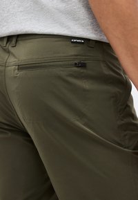 Κοντινό πλάνο χακί παντελονιών με πλαϊνή τσέπη με φερμουάρ και μια μικρή μαύρη ετικέτα πάνω από την τσέπη σε ένα άτομο που φοράει γκρι πουκάμισο.