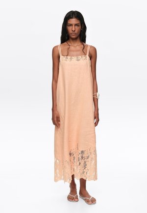 Mujer de pie con vestido melocotón sin mangas con detalles de encaje en el escote y el dobladillo, combinado con sandalias minimalistas y un accesorio de muñeca.
