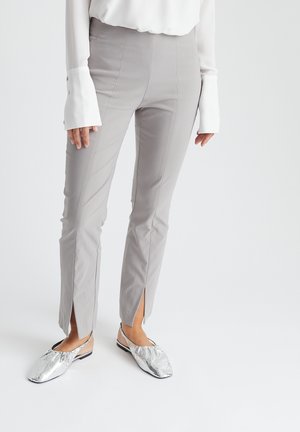 Des pantalons ajustés gris clair présentent une taille haute, une longueur légèrement raccourcie et des fentes sur les côtés à l'ourlet. Associés à des chaussures argentées à enfiler.