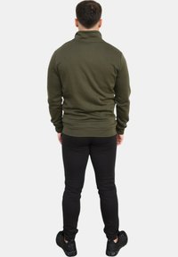 Pullover verde oliva con colletto alto, orlo a coste e texture liscia, abbinato a pantaloni neri aderenti e scarpe da ginnastica nere.