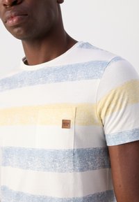 T-shirt kratkih rukava s okruglim izrezom, s horizontalnim prugama u plavoj i žutoj boji na bijeloj podlozi, s malim brendiranim oznakom na prsima.