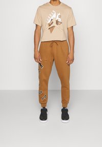 Man i beige Jordan Air t-shirt, bruna joggers med svart och vit text, samt svarta sneakers med vita sulor, står framför en enfärgad bakgrund.