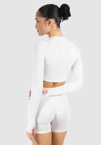 Hvid langærmet crop top med ribbet tekstur og tommelfingerhuller, parret med hvide højttaljede shorts, vist fra en bagvinkel.