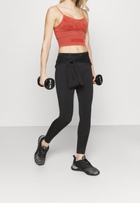 Rød crop top med tynde stropper og et logo, parret med sorte højtaljede leggings og sorte atletiske sneakers, mens der holdes vægte.