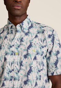 Kortiemige shirt met een blauw bloemenpatroon, voorzien van witte en aqua accenten. De knopen zijn geel en blauw. Inclusief een klein logo op de borst.