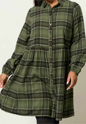 Długa sukienka na guziki w zielono-czarną kratę z marszczoną talią i elastycznymi mankietami, noszona przez osobę trzymającą rąbek.