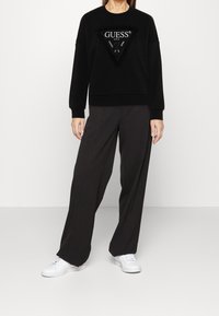 Sudadera de forro polar negra con un parche de logo triangular, combinada con pantalones grises holgados y zapatillas blancas. La tela tiene una textura suave.