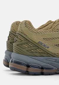 Chaussures de sport vert olive avec tige en mesh, présentant des détails texturés et un rembourrage gris. Marquage distinctif sur le côté. Semelle en caoutchouc.