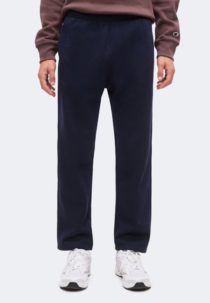 Homme portant un pantalon de survêtement bleu marine associé à des baskets blanches et un haut marron à manches longues, debout devant un fond uni.