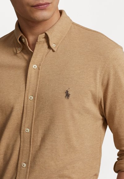 Beige knappskjorte laget av teksturert stoff, med en liten brodert logo på venstre bryst og runde knapper langs plaggåpningen.
