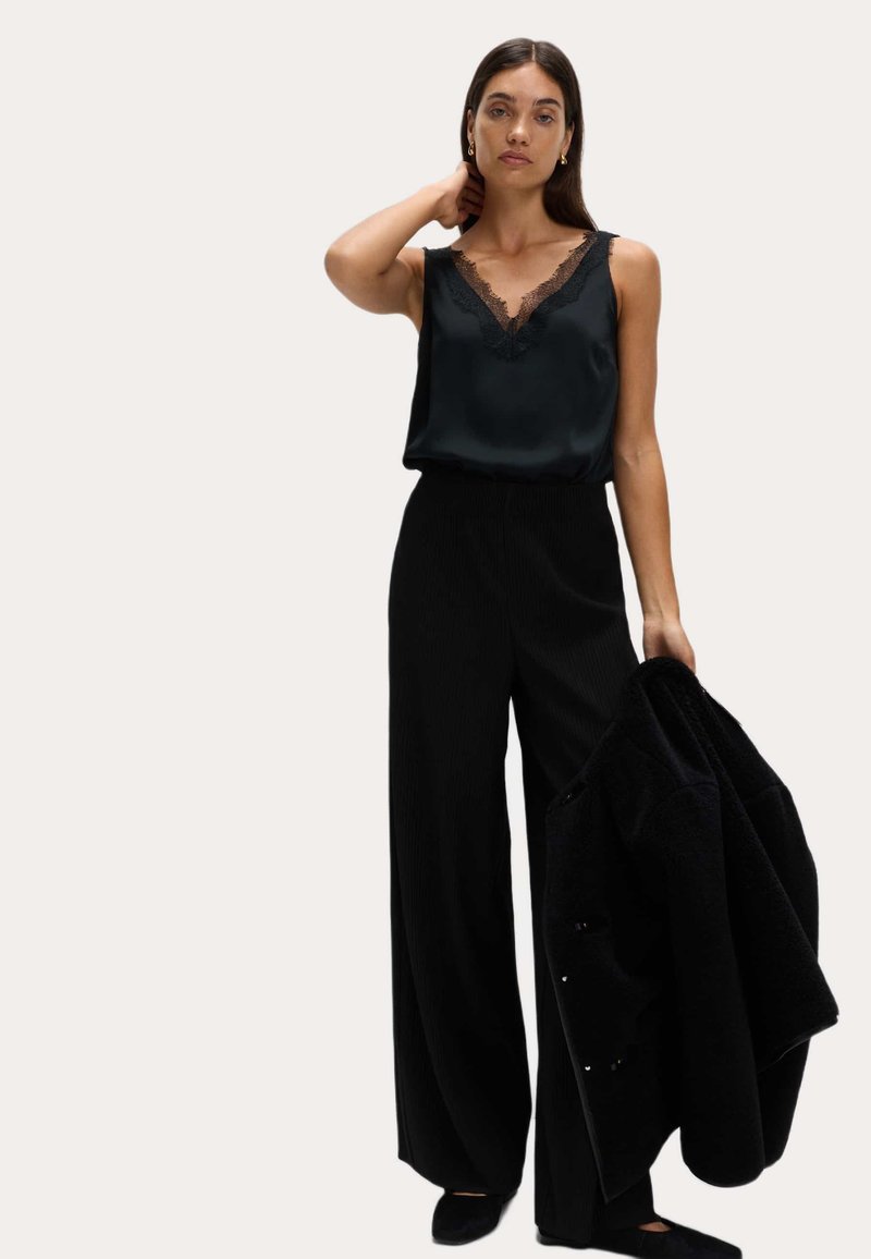 Woman wearing black lace-trimmed sleeveless top and wide-leg pants, holding a black coat in her right hand against a plain background.