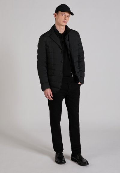 Jonge man met een zwarte pet, een zwart doorgestikt colbert, een zwart overhemd, zwarte broek en zwarte schoenen, staand tegen een lichtgrijze achtergrond.