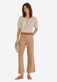 Beige Bluse mit Puffärmeln und Knopfleiste, kombiniert mit hochgeschnittenen, weiten khakifarbenen Hosen und einem braunen Gürtel. Sandalen und eine strukturierte Clutch.