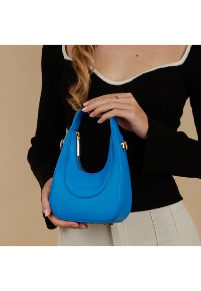 Kleine blauwe handtas gemaakt van glad materiaal met een gebogen ontwerp. Voorzien van gouden hardware en een rits, wordt met één hand vastgehouden.