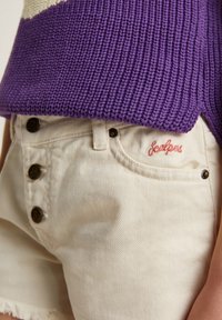 Top de punto morado con textura acanalada, combinado con pantalones cortos de mezclilla de color blanco roto que cuentan con cinco botones de latón y un logotipo bordado en el bolsillo.