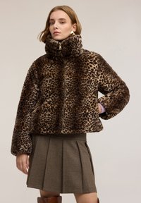 Kobieta ubrana w kurtkę z wysokim kołnierzem zapinaną na zamek, z nadrukiem w panterkę z sztucznego futra, plisowaną spódnicę w kratę oraz duże złote kolczyki koła na jednolitym tle.