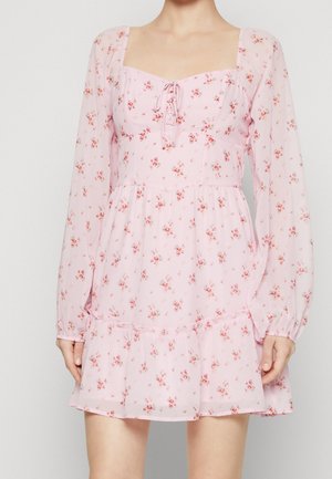 Woman wearing a light pink floral dress with long sheer sleeves, a square neckline, front tie detail, and a ruffled hem.
