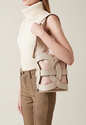 Borsa strutturata beige con design a incrocio in tessuto testurizzato, con dettagli in metallo dorato. Indossata in modo casual con pantaloni marroni a fantasia.