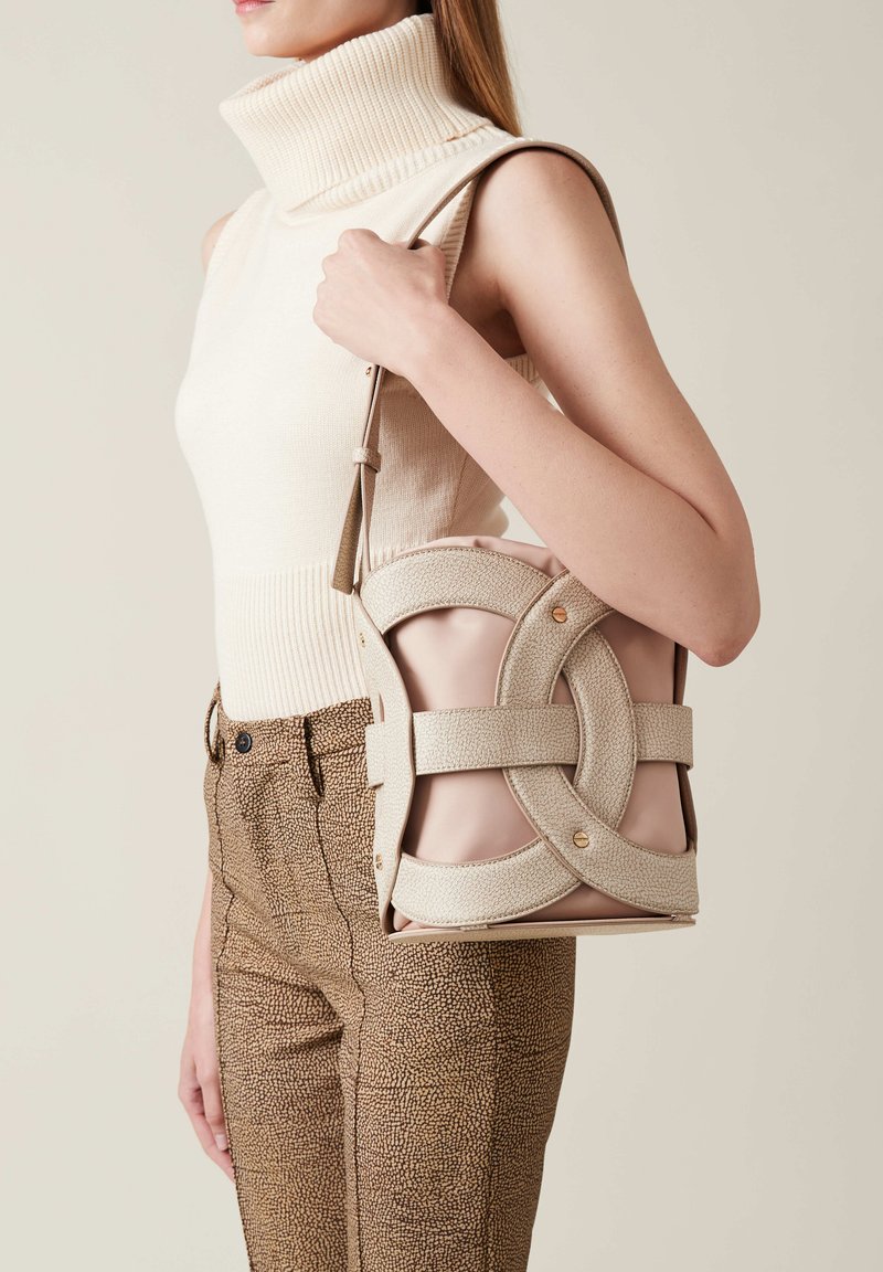 Beige structured handbag with crisscross design in textured fabric, featuring gold hardware accents. Worn casually with patterned brown pants.