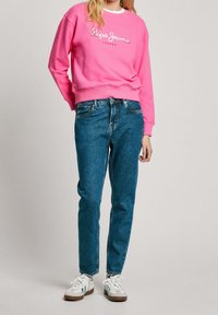 Žena v jasně růžové mikině "Pepe Jeans London", modrých džínách a bílých teniskách stojí před jednobarevným pozadím.