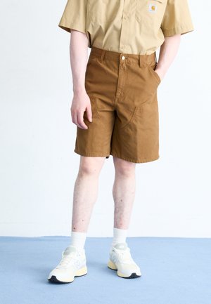 Person trägt ein kurzärmeliges, beige Hemd mit Knöpfen, braune knielange Shorts mit Taschen, weiße Socken und beige Sneakers und steht auf blauem Boden.