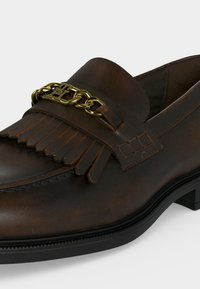 Σκούρο καφέ δερμάτινο loafer με λεπτομέρεια φράντζας και χρυσό αλυσίδα στο πάνω μέρος, με μαύρη σόλα και χαμηλό τακούνι.