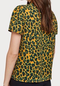 Blouse à manches courtes en tissu jaune moutarde avec un motif léopard vert. Le dos présente une encolure arrondie et une coupe décontractée.