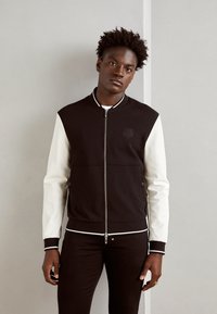 Chaqueta bomber en blanco y negro con cremallera en el frente, mangas contrastantes, puños acanalados y un pequeño logo en el pecho. Tejido suave con una textura lisa.