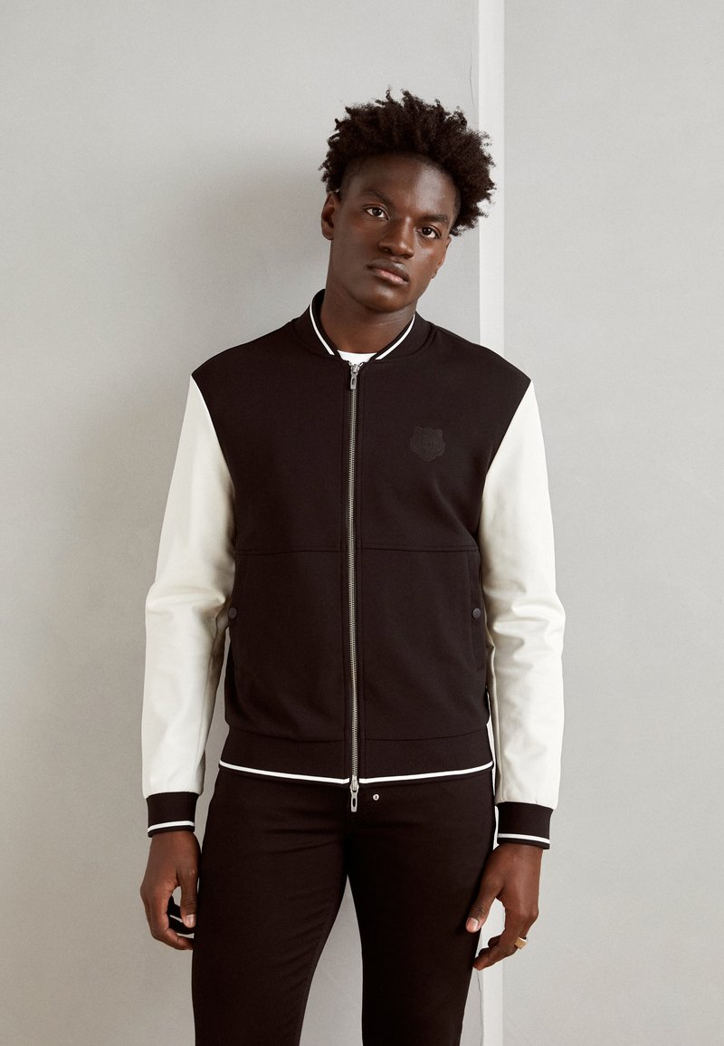 Chaqueta bomber en blanco y negro con cremallera en el frente, mangas contrastantes, puños acanalados y un pequeño logo en el pecho. Tejido suave con una textura lisa.