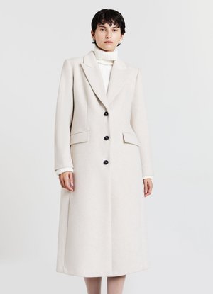 Donna che indossa un lungo cappotto crema con bottoni neri sopra un maglione a collo alto bianco, in piedi davanti a uno sfondo chiaro e semplice.