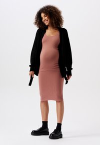 Odzież ciążowa z dopasowaną, prążkowaną różową sukienką oraz czarnym, oversize'owym kardiganem z długimi rękawami i akcentami w postaci wiązań. Całość dopełniają czarne, platformowe buty.