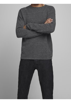Personne debout les bras croisés portant une chemise à manches longues gris foncé et un jean noir sur un fond gris uni.
