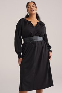 Schwarzes Langarmkleid aus glattem Stoff mit V-Ausschnitt, gerafften Schultern und einem kontrastierenden schwarzen Ledergürtel an der Taille.