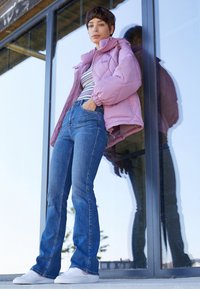 Chaqueta de plumas morada con marca visible, camiseta a rayas, jeans acampanados azules y zapatillas blancas. Reflejo visible en el fondo de cristal.
