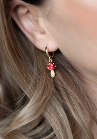 Gold hoop earring with a dangling red mushroom charm, featuring a white spotted cap and a beige stem, textured and playful design.