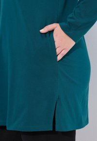 Türkisfarbendes Langarmoberteil mit lockerer Passform, seitlicher Tasche und einem geschlitzten Saum für mehr Bewegungsfreiheit. Glatter Baumwollstoff.