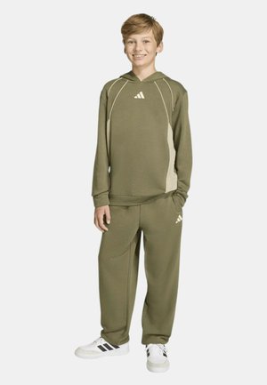 Ragazzo sorridente, indossa una felpa con cappuccio Adidas verde oliva e pantaloni abbinati con sneakers Adidas bianche e nere, in piedi con una mano in tasca.