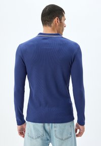 Långärmad, ribbad polo i marinblått, med en klassisk krage med kontrasterande detaljer. Stylen är i kombination med ljusa denimjeans.