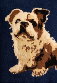 Dekorativt tæppe med en tekstureret blå baggrund og et strikket billede af en brun og cremefarvet hund, der fremhæver detaljerede pelsmønstre.