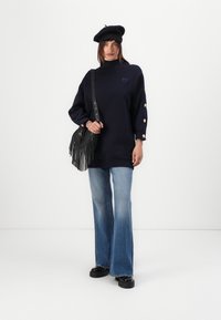 Navy oversized sweatshirt with a high neck, gold button accents on sleeves, paired with flared light blue jeans and a black fringed crossbody bag.