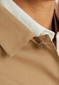 Chemise en tissu beige avec un col cordé blanc, doté d'une fermeture à boutons et de détails de couture visibles, présentant une texture lisse.
