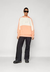 Lekka dwukolorowa bluza z kapturem w kolorach koralowym i kremowym z regulowanym sznurkiem; zestawiona z czarnymi spodniami roboczymi i czarnymi butami.