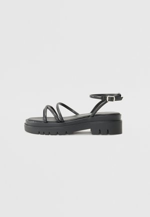 Black strappy sandal with a chunky sole, featuring thin straps and a buckle ankle strap. Smooth texture with a rugged outsole design.