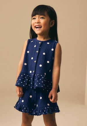 Mosolygó fiatal lány egyenes fekete hajjal, tengerészkék ujjatlan felsőt és fehér pöttyös rövidnadrágot visel, bézs háttér előtt áll.