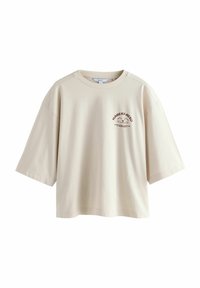 Beige oversized t-shirt i bomull med korta ärmar, med en tryckt logotypdesign i mörkbrunt på framsidan. Rund halsringning, avslappnad passform.