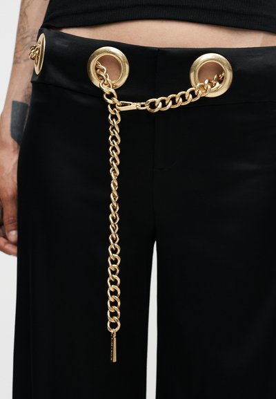 Μαύρο παντελόνι με μεγάλα χρυσά γκρομέτα και χοντρή χρυσή αλυσίδα ζώνης περασμένη μέσα από αυτά, που κρέμεται μπροστά.