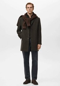 Abrigo largo de lana marrón con cierre de botones, combinado con una camisa oscura y jeans de mezclilla. Bufanda marrón sobre los hombros. Zapatos oscuros.